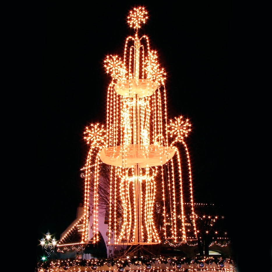 A night scene featuring a towering illuminated display designed to resemble a cascading fountain. The structure is adorned with numerous strings of warm white LED lights, creating a dazzling, festive spectacle. Starburst light patterns accentuate the top and edges of the fountain, adding an extra touch of holiday spirit. The background is dark, making the bright lights stand out vividly against the night sky.