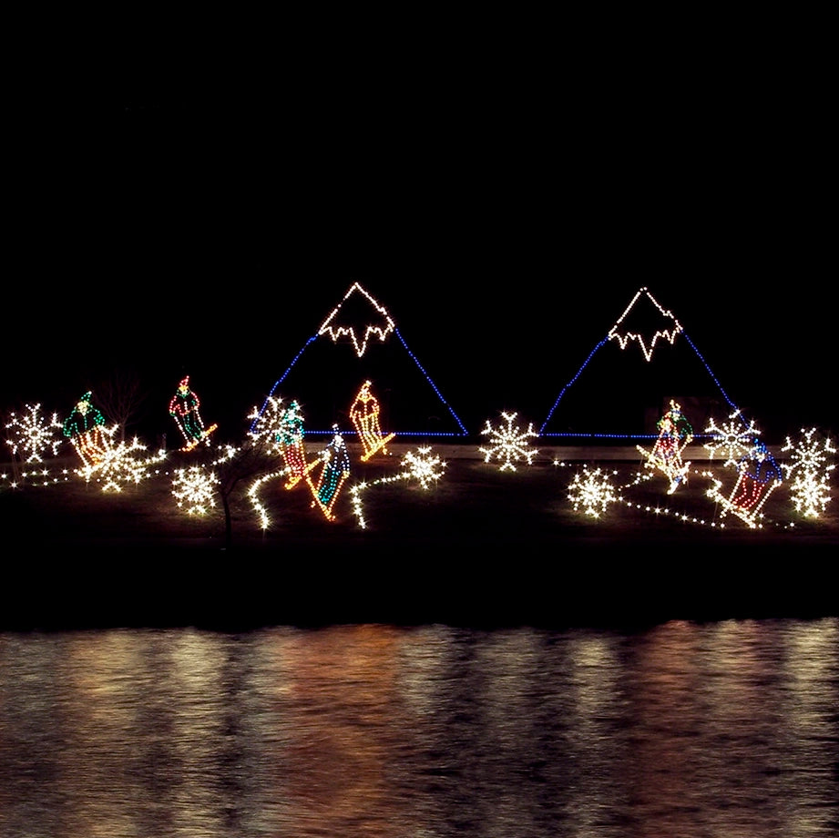 A vibrant night scene featuring a large silhouette LED display. The display depicts a festive winter landscape with snow-covered, purple mountains in the background and several figures skiing down a slope. Bright, colorful lights outline the skiers and create snowflakes scattered across the scene. The display is beautifully reflected in a body of water in the foreground, enhancing the enchanting holiday ambiance.