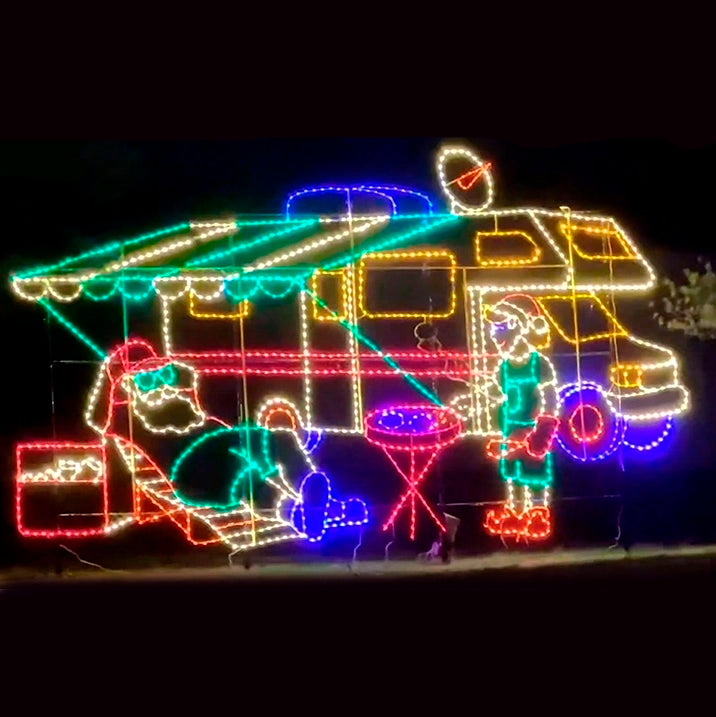 A silhouette LED display featuring Santa Claus, sans Santa hat, relaxing on a lounge chair with sunglasses and a red trucker hat, next to an RV with an awning. A sun elf stands nearby grilling, with vibrant red, green, yellow, white, and blue LED lights outlining the scene, set against a black background.