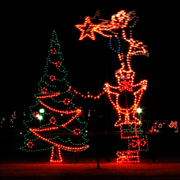 An animated LED silhouette display featuring two bears decorating a large Christmas tree. One bear stands up on stacked Christmas packages holding a string of lights while the other bear balances on the other bear’s head, reaching up to place a star tree topper. The scene is illuminated with bright red, green, blue, and yellow LED lights, set against a dark night background.