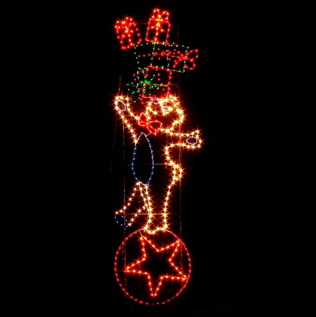 A vibrant silhouette LED display of a Christmas bear balancing on a red ball with a star, holding a stack of brightly wrapped gifts. The display is illuminated with green, yellow, white, red, and blue LED lights, set against a dark background, creating a festive and joyful holiday scene.