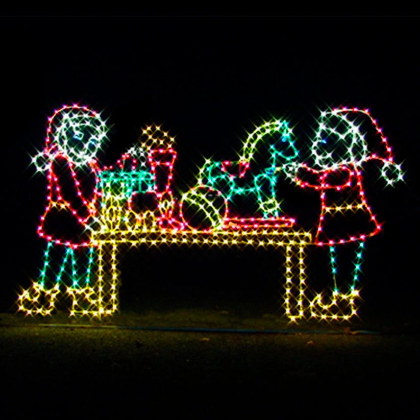 Christmas Elves Making Toys silhouette LED display featuring animated, cheerful elves creating toys. The display is illuminated with bright red, green, blue, and yellow LED lights. The scene shows two elves at a workshop table filled with toys, including a rocking horse and toy train, set against a black background. This vibrant and dynamic display is ideal for businesses, theme parks, municipalities, and event venues looking to enhance their holiday displays.