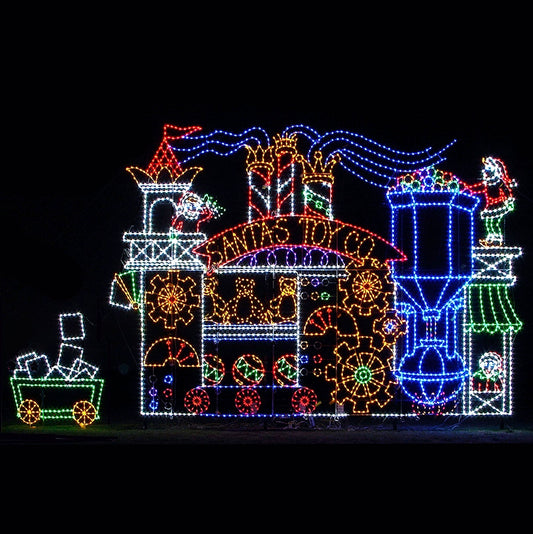 A vibrant and intricate LED silhouette display of "Santa's Toy Company." The display showcases a large toy factory adorned with bright, colorful lights, including spinning gears, candy canes, teddy bears, and festive decor. Santa's elves are busy working, with one loading toys and another managing the machine. The display features animated machine elves with elements and bright colors that illuminate the entire scene against a dark background, capturing the holiday spirit of the Christmas season.