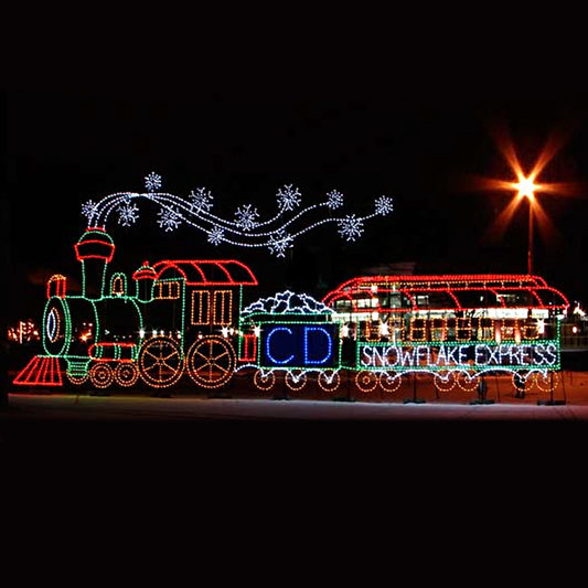 A large, colorful LED display of a train at night, named "Snowflake Express." The train is decorated with bright white, green, blue, and red LED lights. Snowflake patterns are seen emanating from the train's smokestack, adding to the holiday theme. The display is set up in an outdoor public space, illuminated against a dark background.