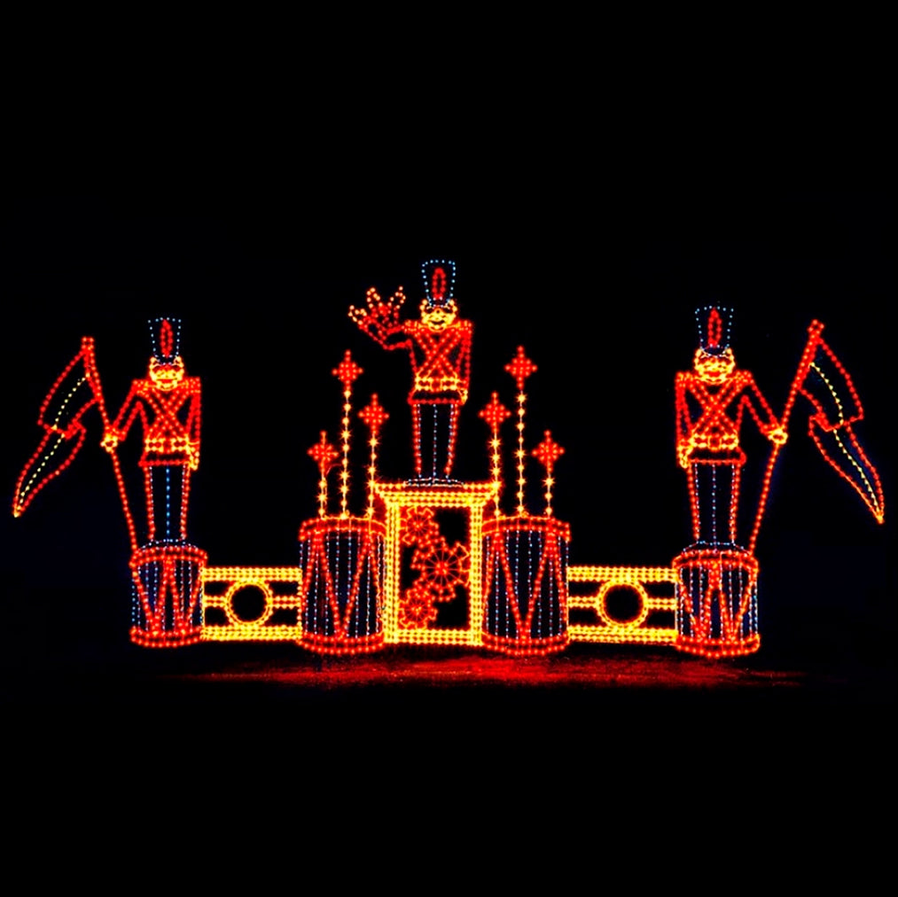 A silhouette LED display featuring three toy soldiers standing on drums, each holding a decorative flag. The soldiers are outlined with bright yellow, blue, and red LED lights, and additional decorative elements are present on the bass drums and snowflake pattern, all set against a black background.