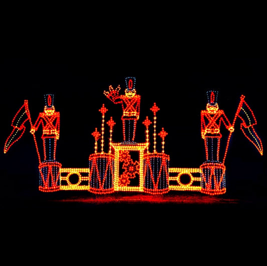 A silhouette LED display featuring three toy soldiers standing on drums, each holding a decorative flag. The soldiers are outlined with bright yellow, blue, and red LED lights, and additional decorative elements are present on the bass drums and snowflake pattern, all set against a black background.